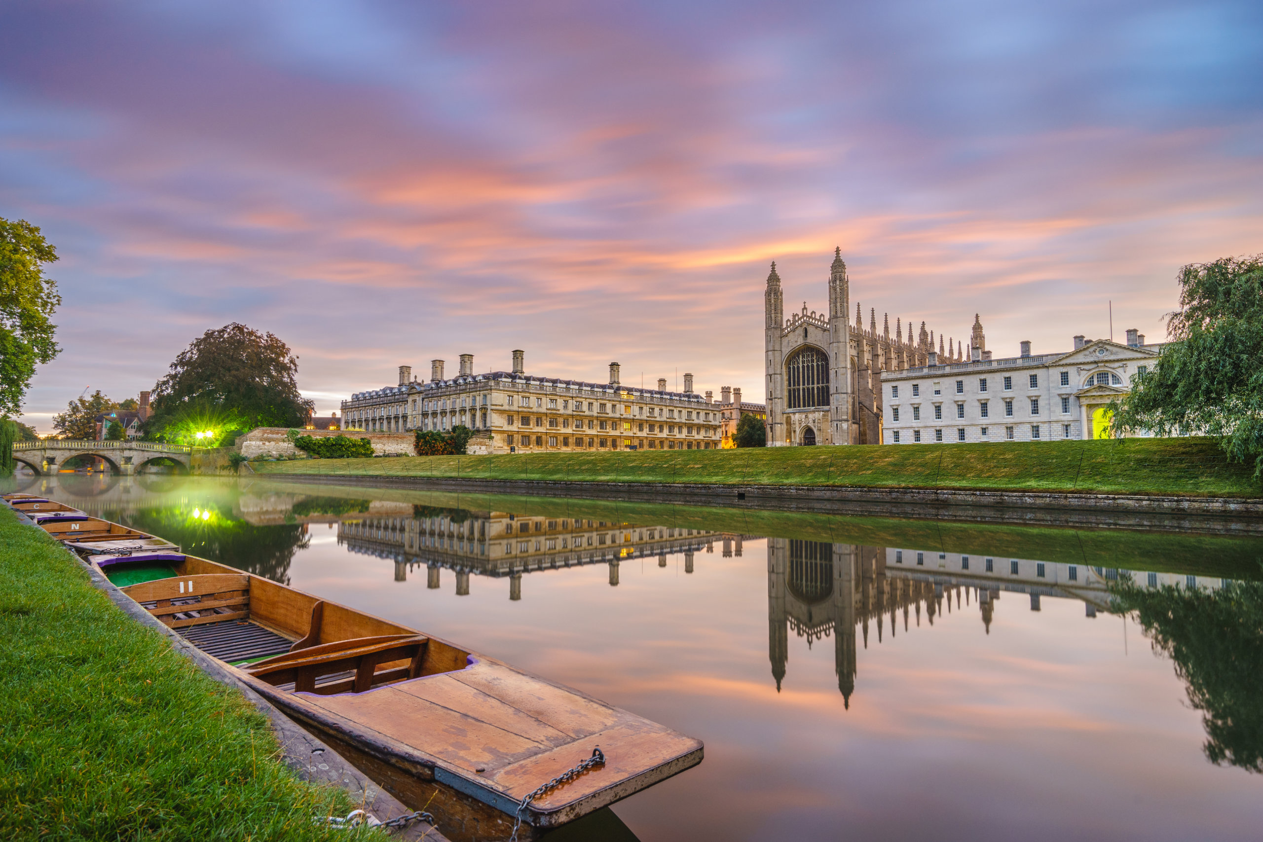A sleeping punt is moored opposite King's College Chapel - one of the beautiful sights that you'll see on your private punting tour. The sky glows pink and the water is serene, still and like a mirror. There's a clear reflection of Clare College and the Chapel. with a the Green lights of a party on Clare Bridge.